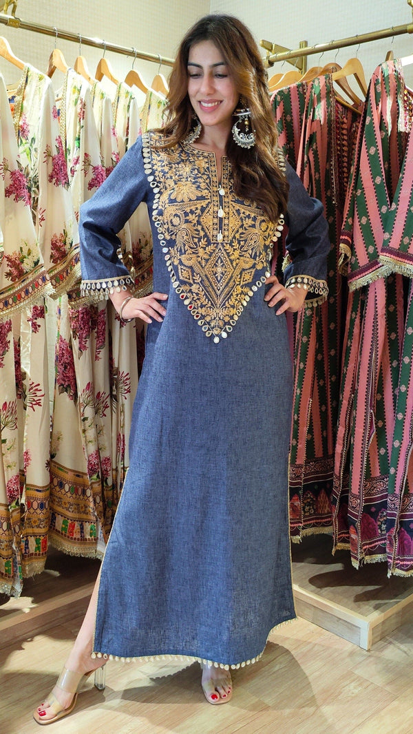 Woman in a blue embroidered dress standing in a clothing store with racks of dresses in the background.