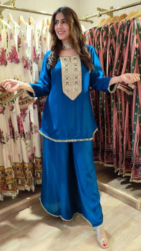 Woman in a blue traditional outfit standing in a clothing store with racks of dresses in the background.