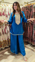 Woman in a blue traditional outfit standing in a clothing store with racks of dresses in the background.